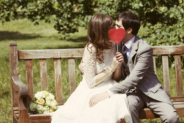 Bride and groom kissing on a bench with their faces covered by a heart - Picture by Rebecca Wedding Photography