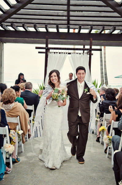 Bride and groom walking from ceremony with Chuppah wrapped around them - Picture by Captured by Aimee