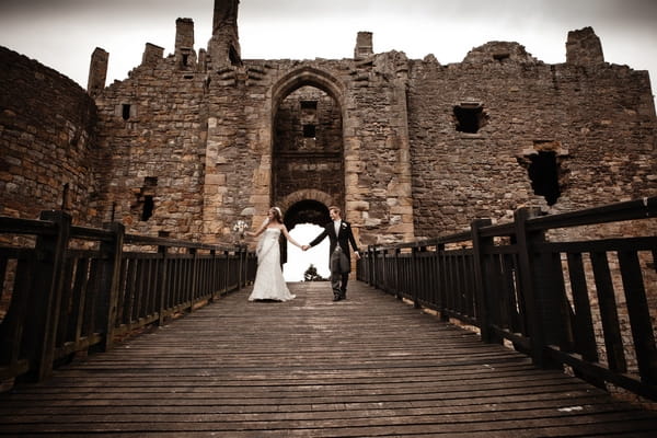 Bride and groom holding hands as they walk across bridge at Dirleton Castle - Picture by Archibald Photography