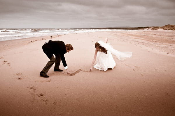 Bride and groom drawing heart in the sand - Picture by Archibald Photography