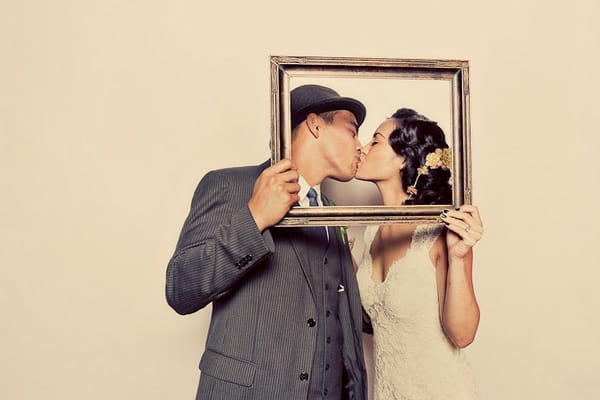 Bride and groom kissing behind picture frame in funbooth - Picture by Paco and Betty