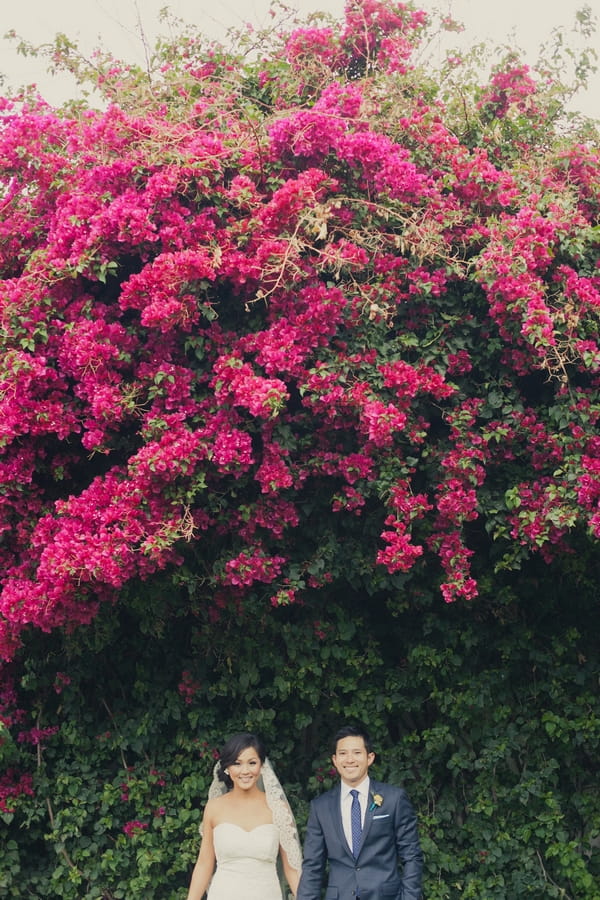 Bride and groom standing under pink flowered tree - Picture by onelove photography