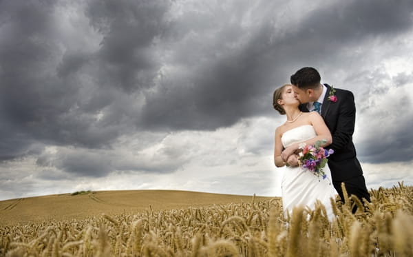 Bride and groom kissing in a field with dark clouds overhead - Picture by Charlotte Snowden Photography