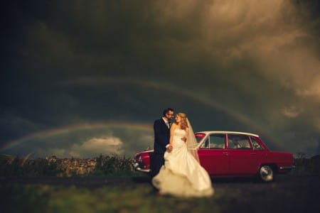 Bride and groom standing by car with dark clouds and rainbow in background - Picture by DSB Creative