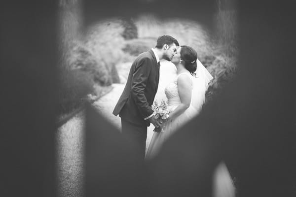 Bride and groom seen kissing through heart shape in fence