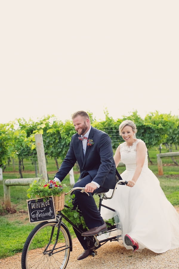 Bride and groom on tandem bicycle cycling past Keswick Vineyards