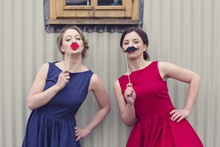 Brides wearing vintage red and blue wedding dresses holding lips and moustache to faces