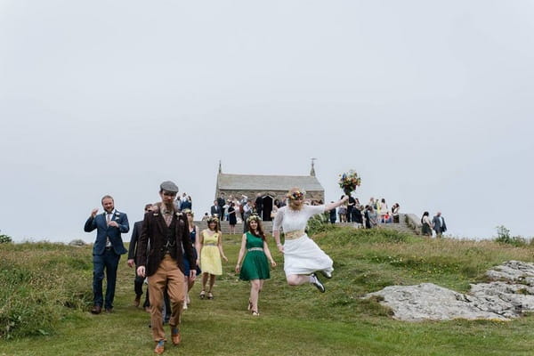Bride jumping in air and clicking heels together as wedding guests walk down hill - Picture by Alexa Loy Photography
