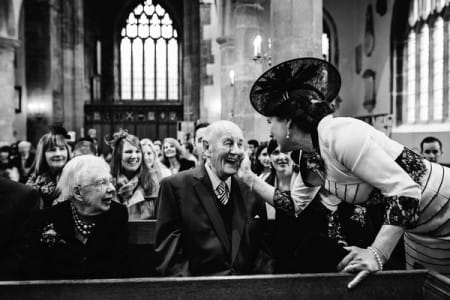 Lady wedding guest touching smiling elderly man's face before wedding ceremony - Picture by How Photography