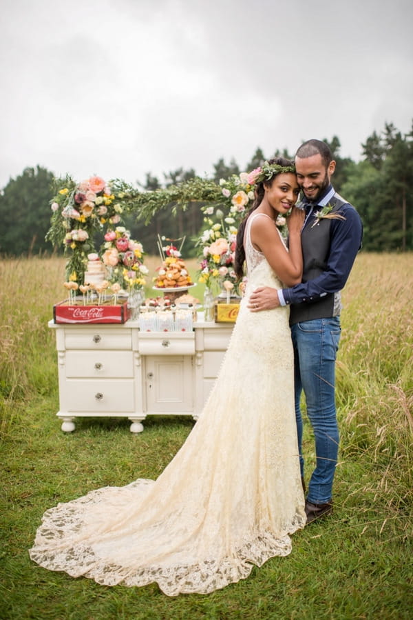 Bride and groom standing in meadow in front of rustic dresser of wedding cakes and desserts