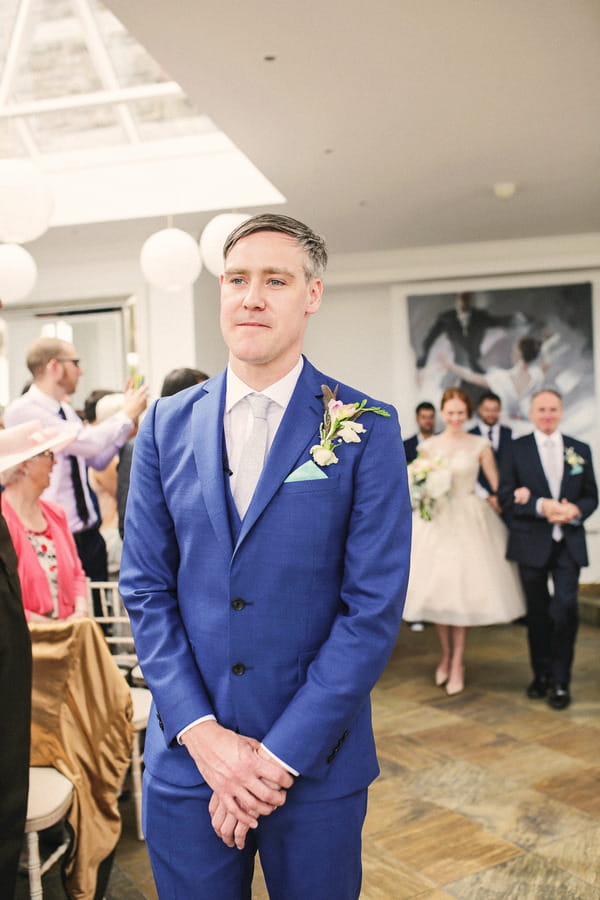 Groom in blue suit with bride walking down aisle behind him