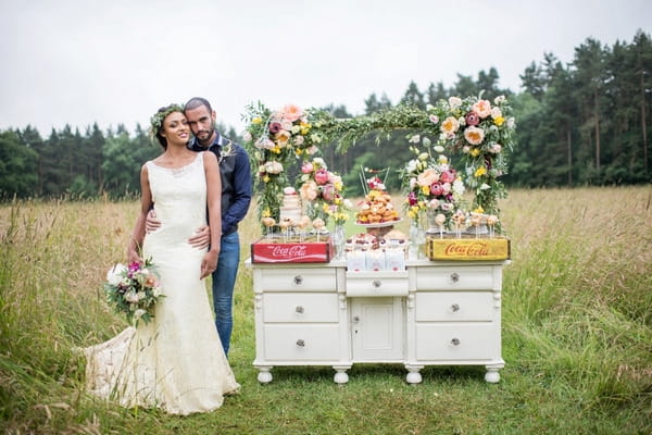 Bride and groom standing in meadow by rustic dresser of wedding cakes and desserts
