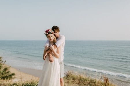 Groom with arms around bohemian bride's shoulders on beach in Spain