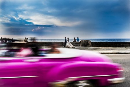 Bride and groom walking across seafront as pink car goes past in a blur - Picture by Augustin Regidor