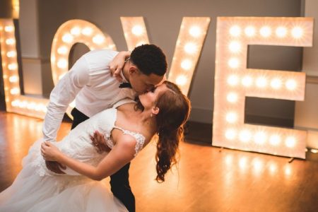 Bride and groom leaning back kiss with large illuminate LOVE letters behind - Picture by Makowski Photography