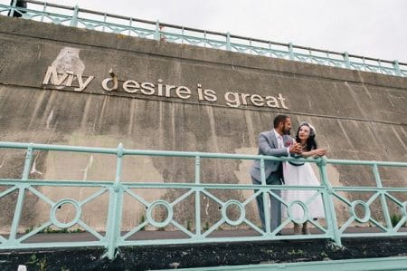 Bride and groom leaning on railings in front of lettering which reads My Desire is Great - Picture by Eclection Photography