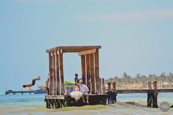 Bride and groom kissing on end of jetty as man jumps off