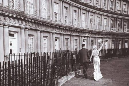 Bride holding bouquet in air as she walks past row of houses with groom - Picture by Steph Newton Photography