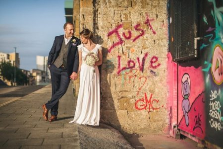 Bride and groom leaning against wall with Just Love written in graffiti - Picture by Andy Hudson Photography