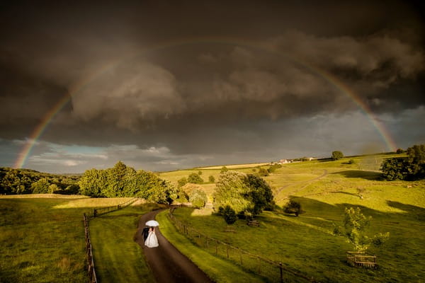 Bride and groom walking down road in beautiful countryside with darks clouds and rainbow overhead - Picture by Ian Baker Photography