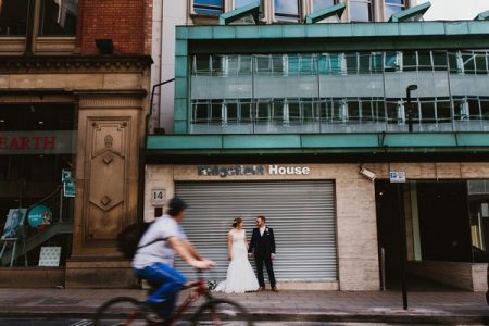 Bride and groom standing in front of shop shutter as cyclist rides by - Picture by Ally M Photography