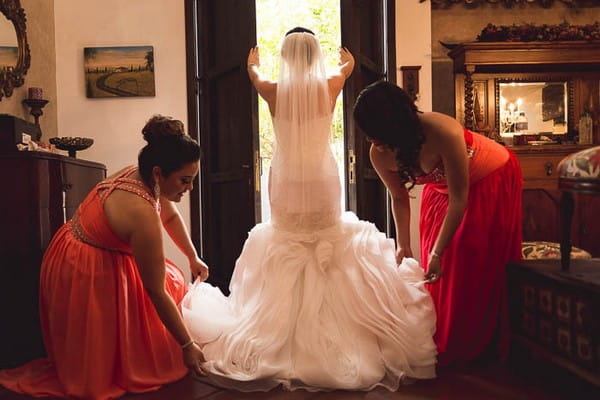 Bride standing at window as bridesmaids pull out train of her dress