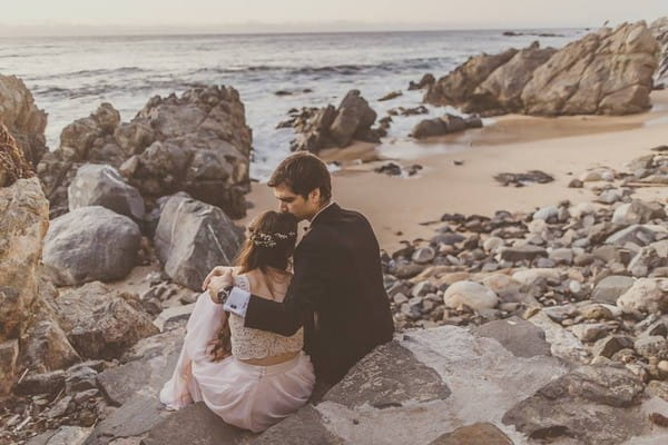 Groom kissing bride's head as they sit on rocks by the sea - Picture by Michelle Lindsell Photography