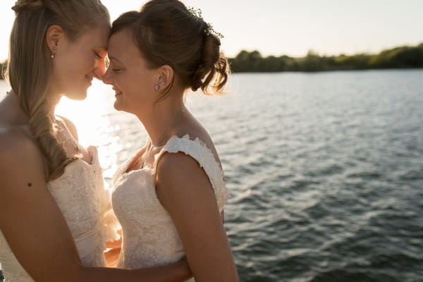 Two brides touching heads in front of water - Picture by Becky Harley Photography