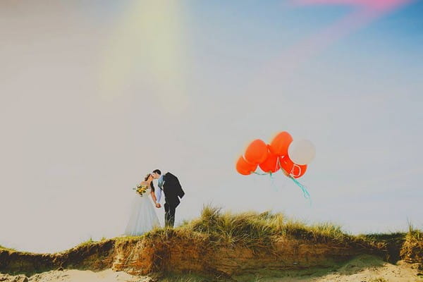 Bride and groom kissing on top of a hill next to orange balloons - Picture by Silver Birch Photography