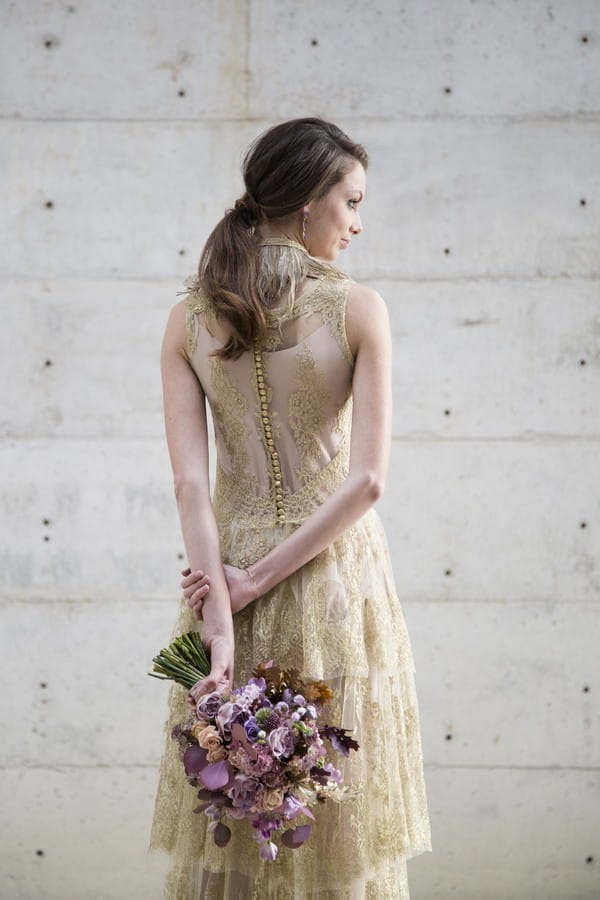 Bride in gold wedding dress holding purple bouquet behind her back