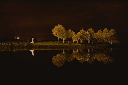 Bride and groom standing by lake at night with trees reflecting in the water - Picture by Lee Allison Photography