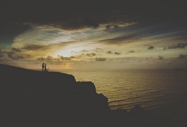 Bride and groom on cliff in front of sea with dramatic clouds overhead - Picture by Howelljones Photography
