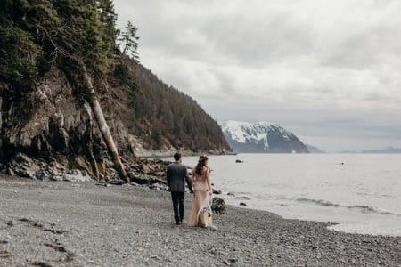 Bride and groom walking across pebble beach with mountain in background - Picture by Kristian Irey Photography