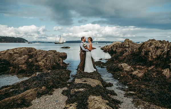 Bride and groom standing on rocks by sea - Picture by Tracey Warbey Photography