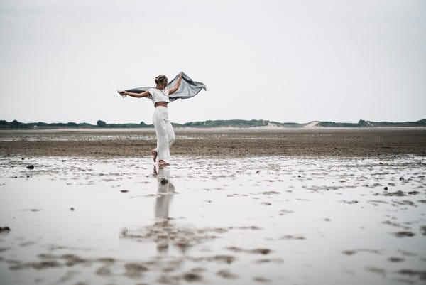 Bride wearing bridal trousers and top holding out shawl on beach