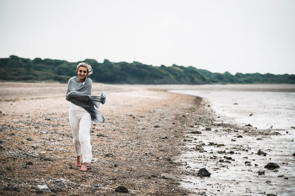 Bride wearing bridal trousers and top walking on beach with shawl wrapped around her