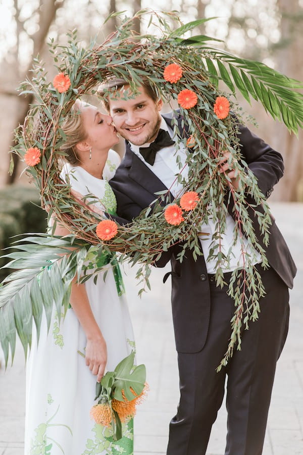 Bride kissing groom behind wreath of orange flowers