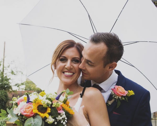 Smiling bride holding umbrella with groom about kiss her on cheek - Picture by Rebecca Gurden Photography