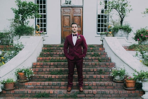 Groom in burgundy suit on steps of Natte Valleij