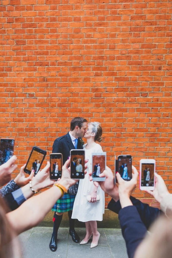 Guests taking pictures on their phones of bride and groom kissing