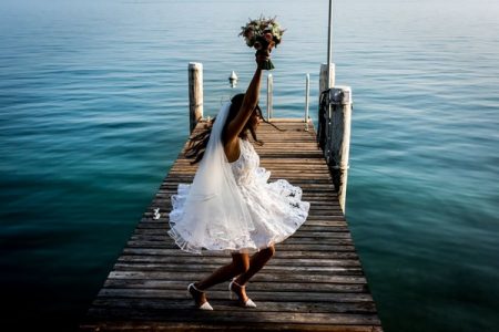 Bride in short wedding dress holding bouquet abover her head on jetty - Picture by Carine Bea