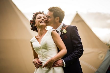 Groom kissing bride on cheek from behind in front of tipis - Picture by Linus Moran Photography