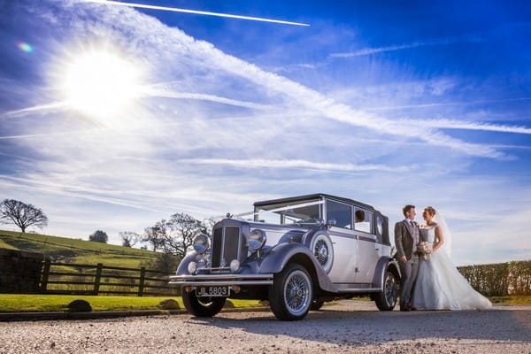 Bride and groom standing by wedding car with beautiful blue sky overhead - Picture by SJPhotographers