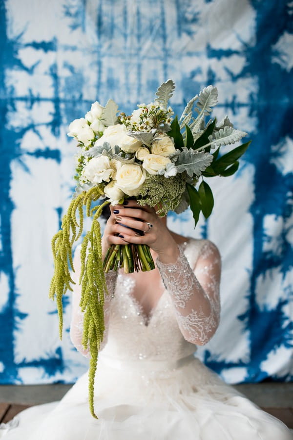 Bride holding wedding bouquet of white flowers and green foliage in front of her face