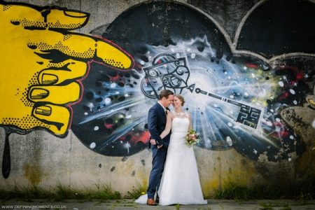 Bride and groom standing against wall with a graffiti hand pointing at them - Picture by Defining Moments Photography