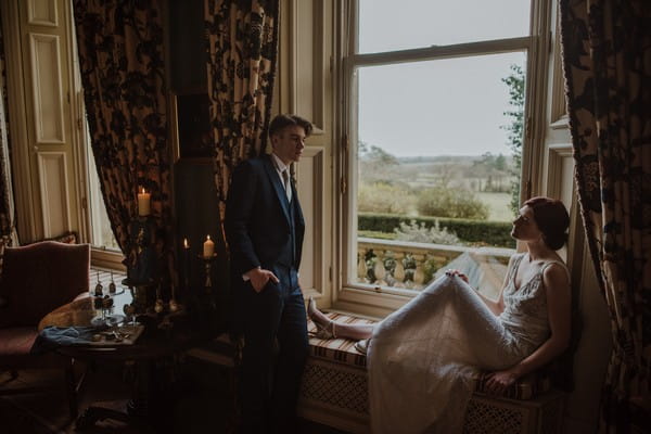 Groom standing by bride sitting on window ledge