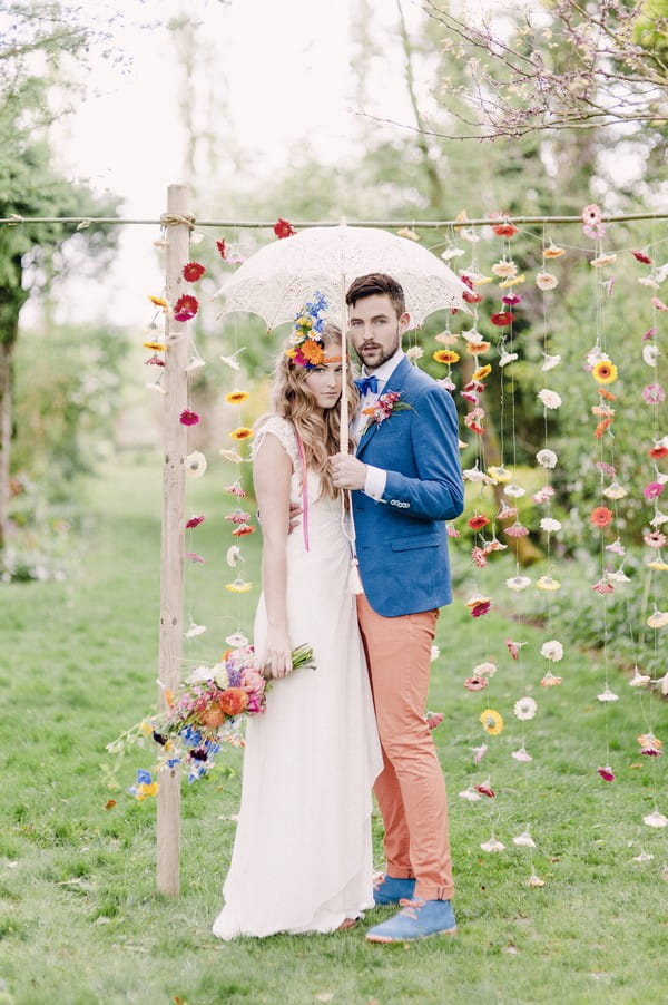 Bride and groom with parasol standing in front of colourful suspended floral backdrop