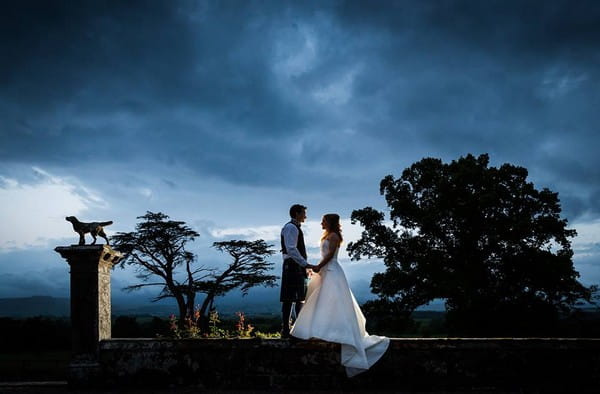 Bride and groom standing on a wall holding hands - Picture by First Light Photography