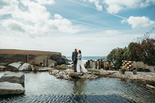 Bride and groom standing on rocks by pool of water - Picture by Tracey Warbey Photography