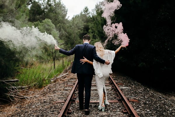 Bride and groom holding smoke bombs walking down train track